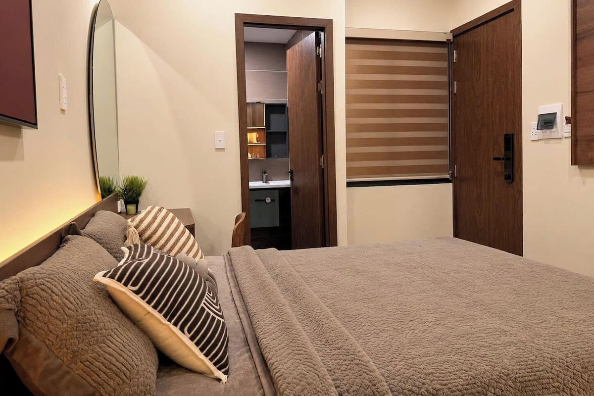 A bedroom with a bed, a bedside table, a mirror, a wardrobe, and a bathroom visible through an open door.