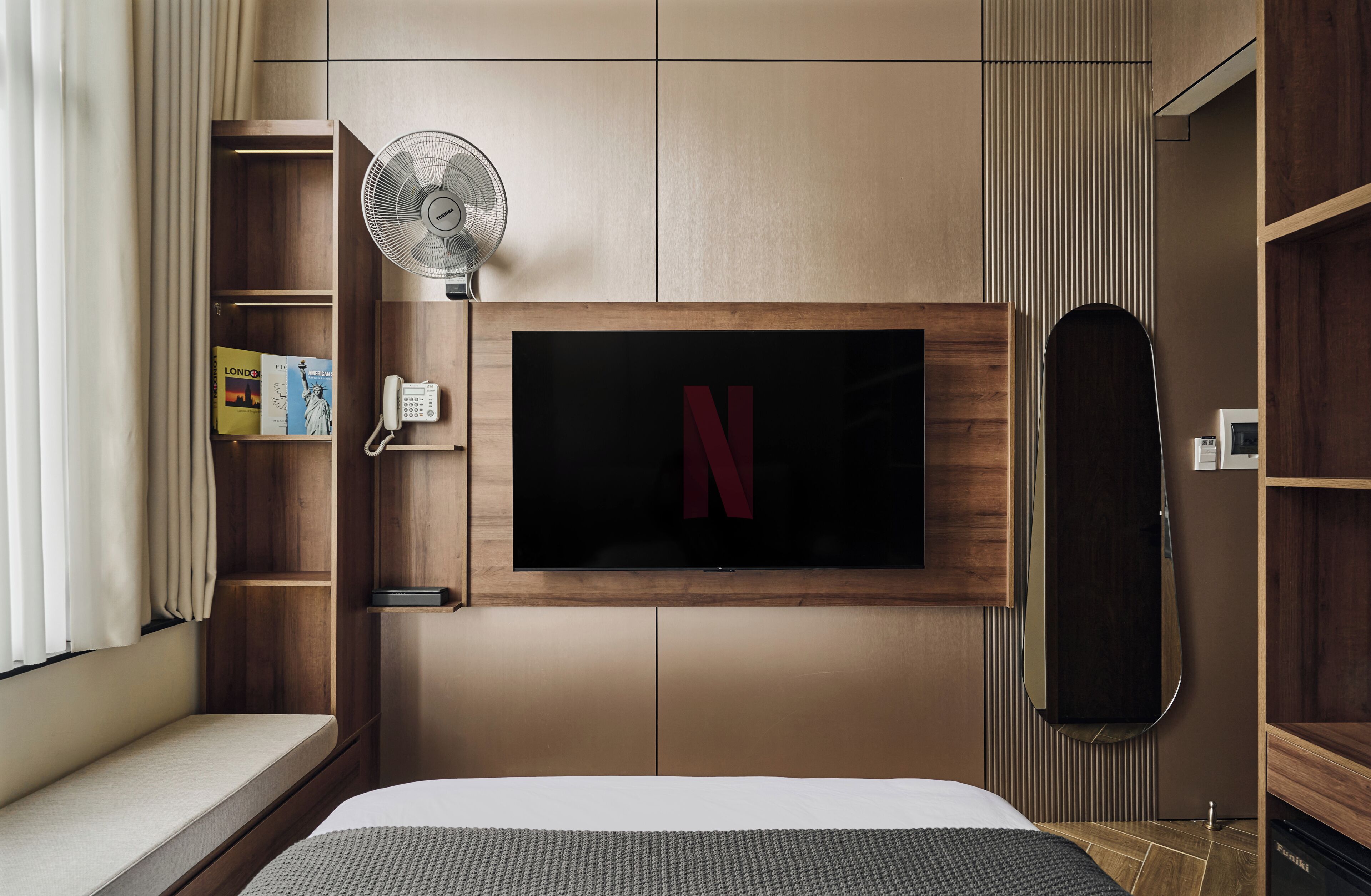 Une chambre d’hôtel avec un lit, un téléviseur fixé au mur, un ventilateur et une étagère en bois avec des livres.