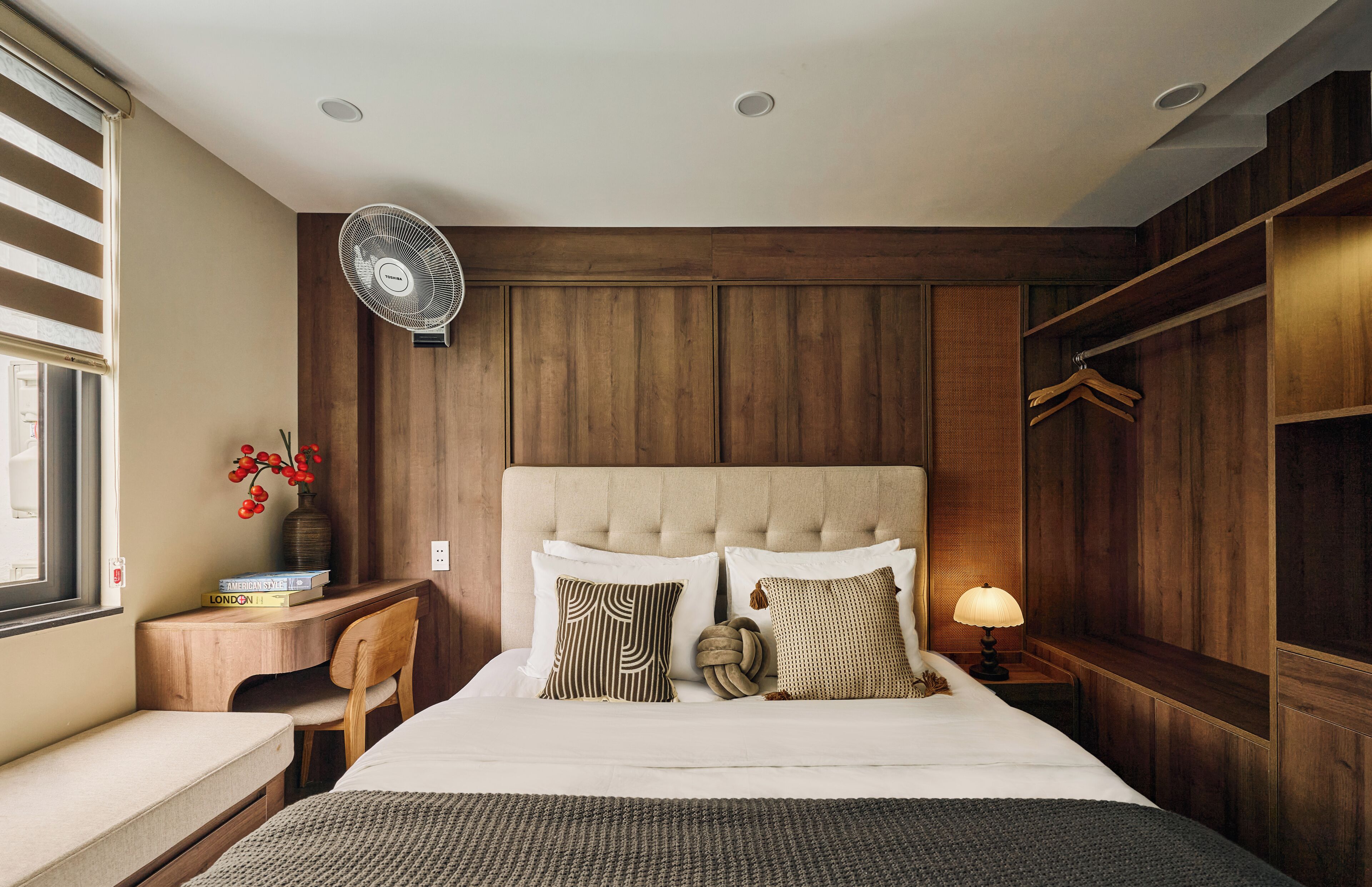 Un dormitorio con cama, mesita de noche, silla, lámpara y ventilador de pared.