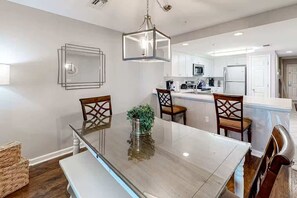 A modern kitchen with white cabinets, a central island, and a dining area with a glass table and wooden chairs. - Amalfi D327 - 3rd Floor Views & Cabana Included! (Miramar Beach)