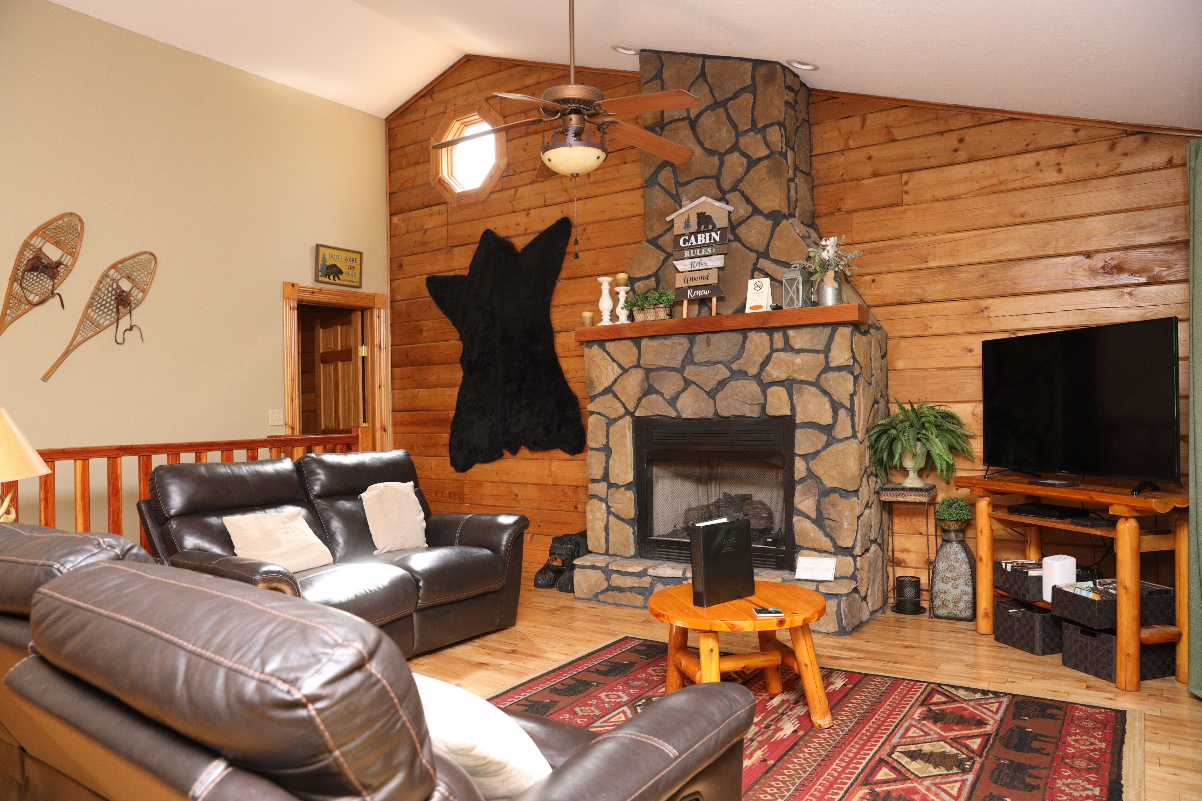 Un salon chaleureux, aménagé dans le style d’un chalet, avec une cheminée en pierre, des canapés en cuir et un téléviseur à écran plat.