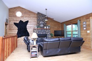 A cabin interior with a stone fireplace, a leather sofa, a television, and a wooden ceiling.