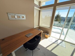 Een modern kantoor met een houten bureau, een zwarte bureaustoel en een groot raam met uitzicht op een zwembad en bomen.