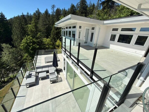 Ein modernes Haus mit einem Glasbalkon, Sitzgelegenheiten im Freien und Blick auf ein bewaldetes Gebiet.
