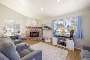 A living room with a fireplace, a clock, a bear painting, and a large window. - Relaxing Cabin- Hot Tub- Lake View-Fire Pit-Hiking- Walk to Restaurants, Shops (Big Bear Lake)