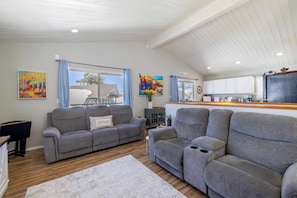 A living room with two grey sofas, a coffee table, and a rug. There are windows with curtains, a painting, and a kitchen area with cabinets. - Relaxing Cabin- Hot Tub- Lake View-Fire Pit-Hiking- Walk to Restaurants, Shops (Big Bear Lake)