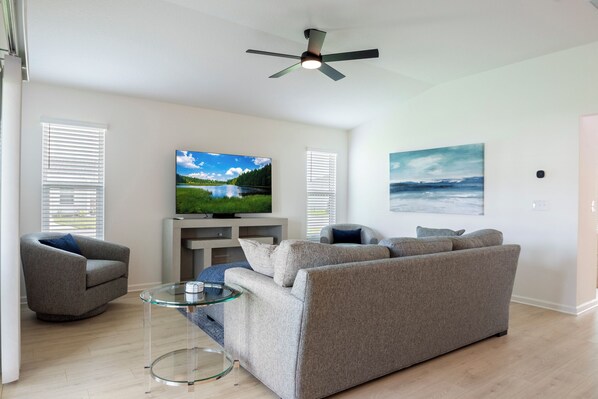 Ein Wohnzimmer mit einer Couch, Sesseln, einem Glastisch, einem Deckenventilator, einem Fernseher und Kunstwerken an den Wänden.