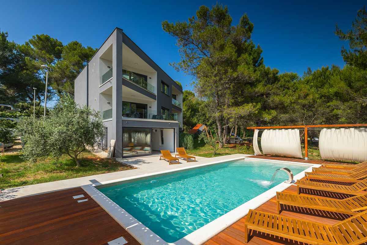 A modern two-story building with a swimming pool, wooden deck, and lounge chairs.