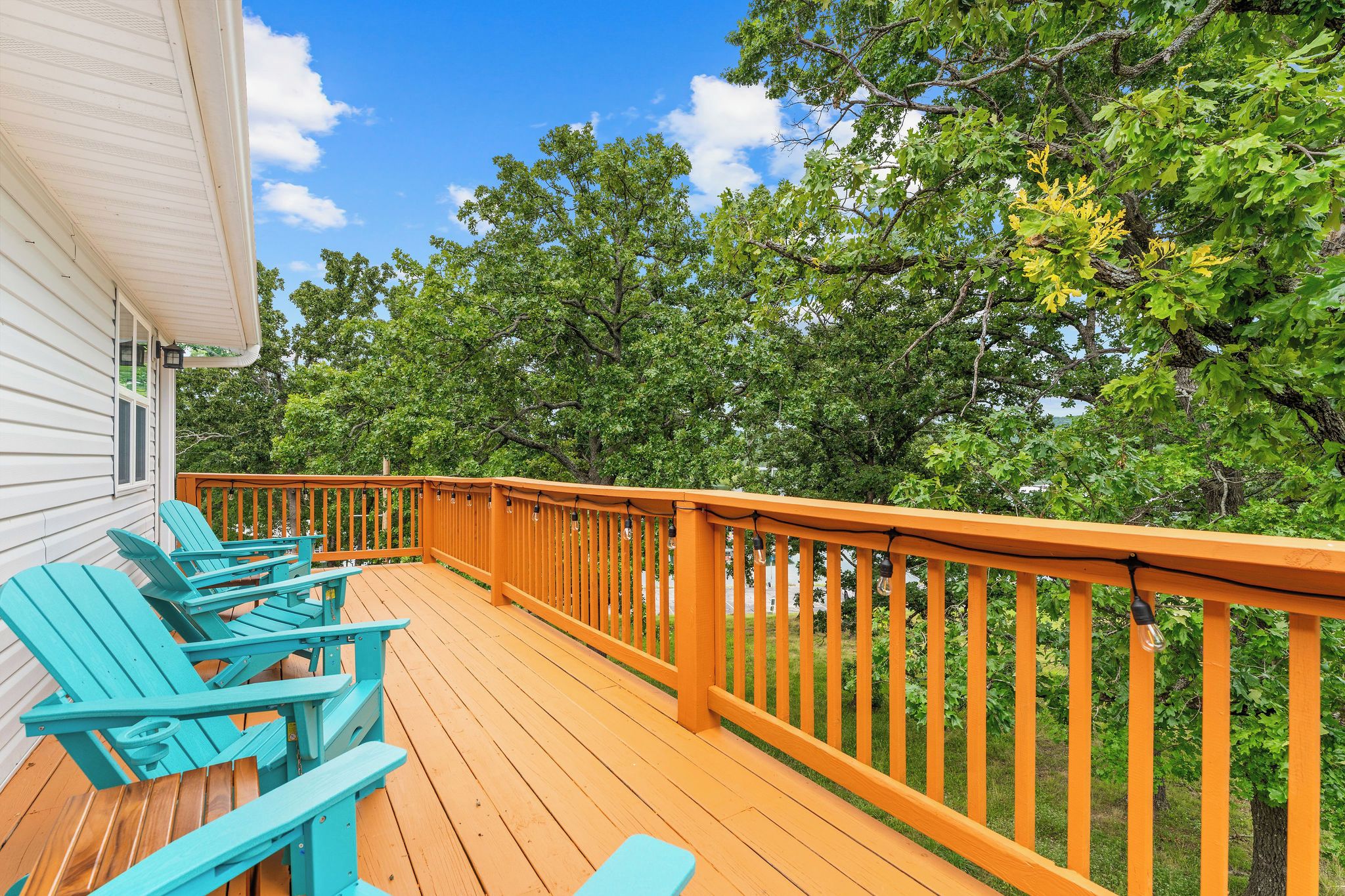 A wooden deck with a railing, turquoise Adirondack chairs, and a view of trees.