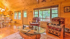 A wooden cabin interior with a glass coffee table, two patterned armchairs, a dining area, and large windows.