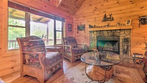 A cozy cabin interior with wooden walls, a stone fireplace, two patterned armchairs, and a glass coffee table.