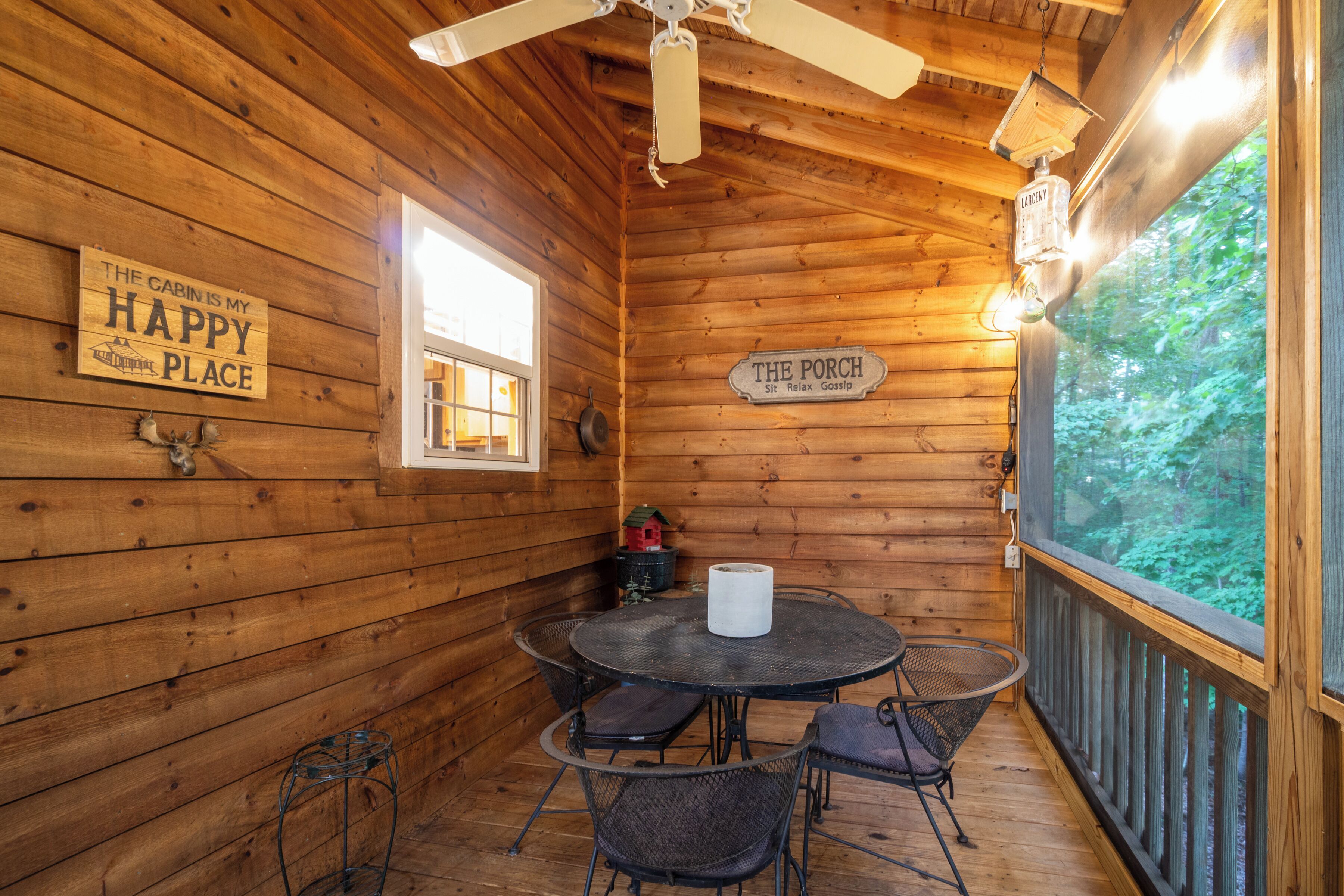 Interior de una cabaña de madera con una mesa redonda y sillas, un ventilador de techo y carteles que dicen 'The Cabin is My Happy Place' y 'The Porch'.