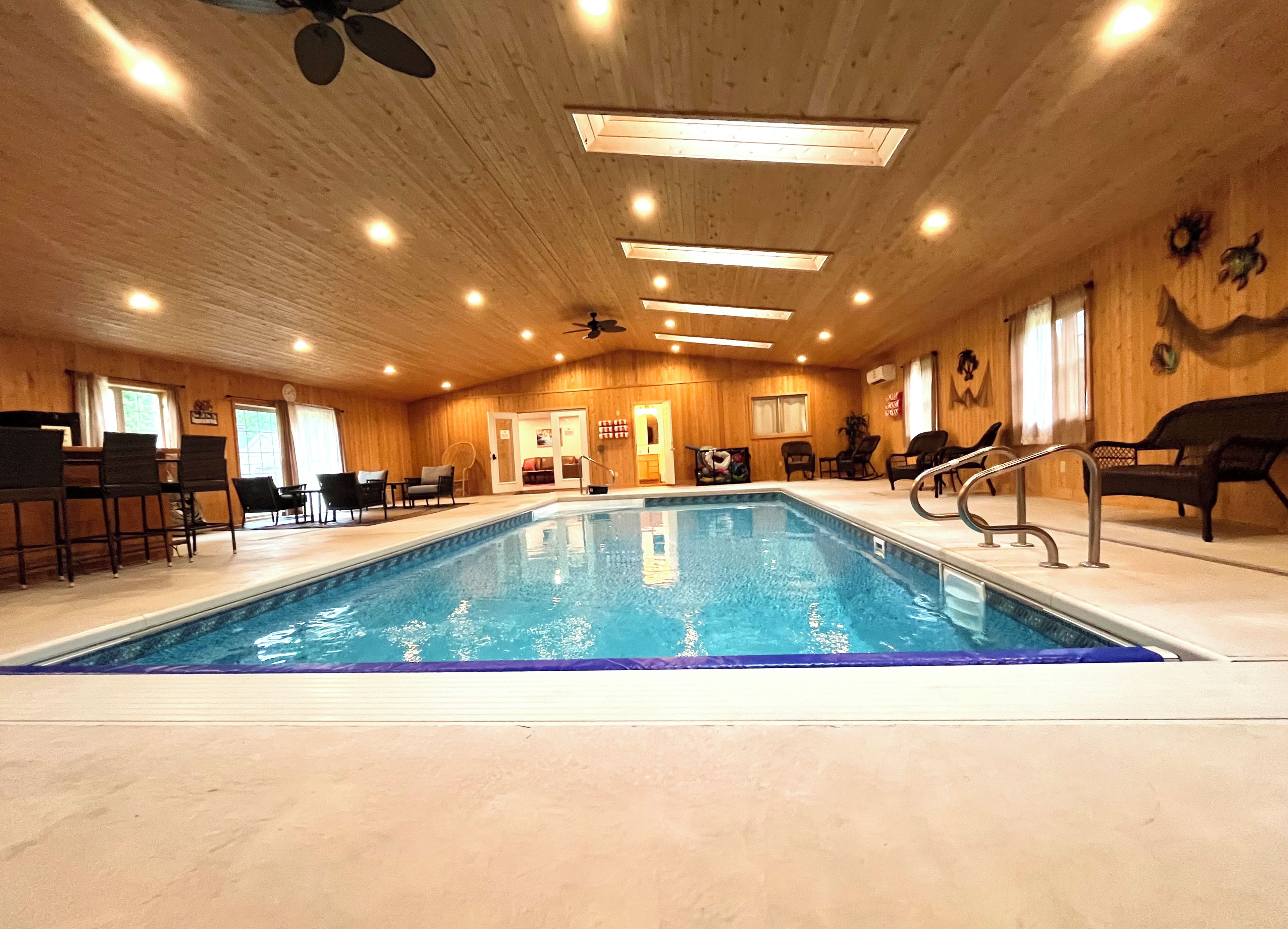 An indoor swimming pool with a wooden interior, surrounded by lounge chairs and a ceiling fan.