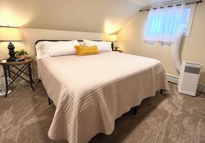 A neatly made bed with a quilted bedspread, two bedside lamps, a small potted plant, and a window with a white curtain.