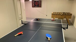Ein Tischtennistisch mit zwei Schlägern, eine Dartscheibe und ein Kicker in einem Raum mit weißen Wänden.