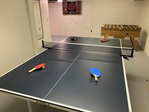 A ping pong table with two paddles, a dartboard, and a foosball table in a room with white walls. - Indoor Heated Pool inside Spacious 4-bedroom house in serene Escanaba with AC (Escanaba)