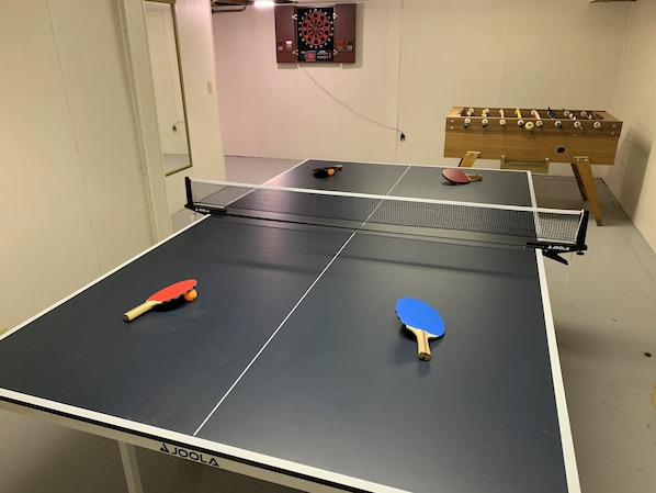 Una mesa de ping pong con dos paletas, un dardo y una mesa de futbolín en una habitación con paredes blancas.