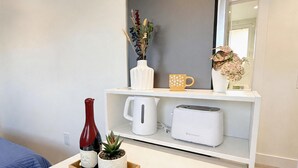 A hotel room with a white bed, a white coffee table with a tray holding a wine bottle and a plant, a white shelf with a coffee mug, a white kettle, and a toaster.