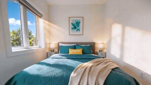 Quarto com cama, janela, travesseiro de boas-vindas e uma obra de arte emoldurada na parede.