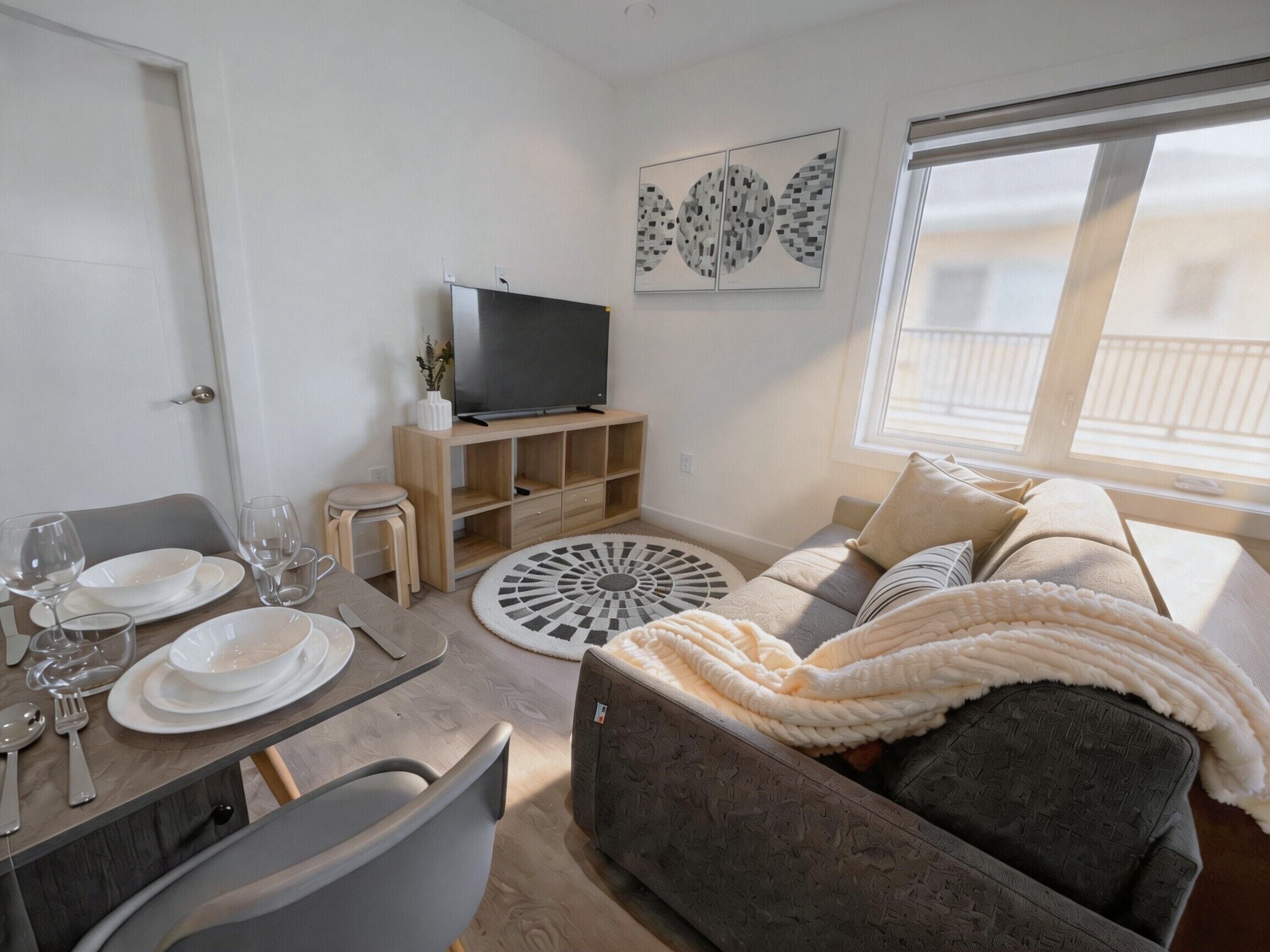 Ein modernes Wohnzimmer mit einer Couch, einem Fernseher auf einem Holzstandfuß, einem Esstisch mit Tellern und Gläsern sowie einem gemusterten Teppich.