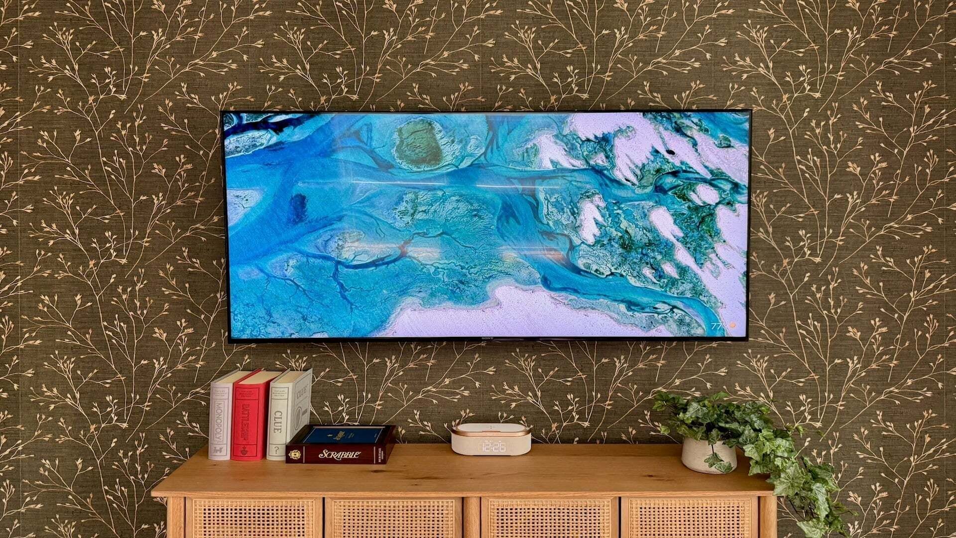 A flat-screen TV mounted on a wall with a patterned wallpaper, a wooden console table with books and a clock, and a potted plant.