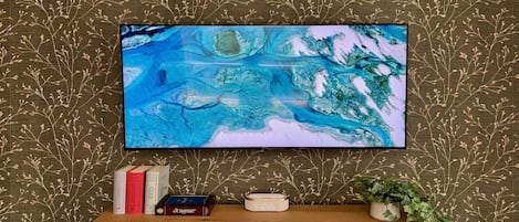 A flat-screen TV mounted on a wall with a patterned wallpaper, a wooden console table with books and a clock, and a potted plant.