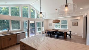 A modern kitchen with a marble countertop, a dining area with a wooden table and chairs, and large windows offering views of greenery. - Woodland - Stunning 5 bedroom home in GB! (Great Barrington)