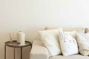 Une pièce aux couleurs neutres, avec un canapé beige, une petite table ronde sur laquelle se trouvent une bougie blanche et un diffuseur de parfum à bâtonnets, et un mur sobre.