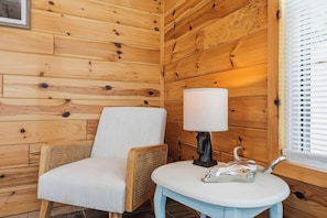Ein Holzblockhaus-Interieur mit einem Korbsessel, einem kleinen Tisch mit Lampe und einem Fenster mit Jalousien.