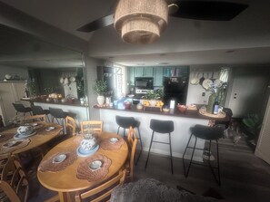 Ein Essbereich mit einem Holztisch, gedeckt für vier Personen, umgeben von Stühlen, und eine Küchenzeile mit Kochinsel, Geräten und verschiedenen Gegenständen auf den Arbeitsflächen.