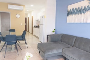 Ein modernes Wohnzimmer mit grauem Sofa, einem Esstisch mit Stühlen, einer Küchenzeile und einer Wanduhr.
