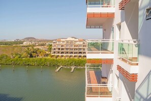 Ein modernes Hotel mit mehreren Balkonen mit Blick auf ein Gewässer und ein in der Ferne befindliches Gebäudeensemble.
