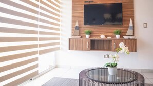Een moderne woonkamer met een aan een houten muur bevestigde televisie, een ronde metalen tafel met een potplant, en een raam met horizontale zonwering.