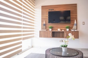 Ein modernes Wohnzimmer mit an einer Holzwand montiertem Fernseher, einem runden Metalltisch mit einem Blumentopf und einem Fenster mit horizontalen Rollläden.