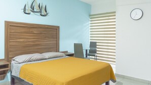A bedroom with a wooden headboard, a bed with a yellow blanket, a nightstand, a chair, a wall clock, and a window with blinds. - Departamento Marina · Alberca · Balcón · Cerritos (Mazatlán)