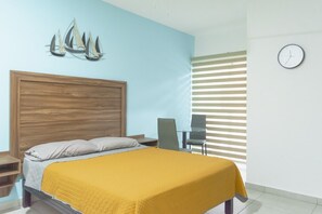 Ein Schlafzimmer mit einem hölzernen Kopfteil, einem Bett mit gelber Decke, einem Nachttisch, einem Sessel, einer Wanduhr und einem Fenster mit Jalousien.