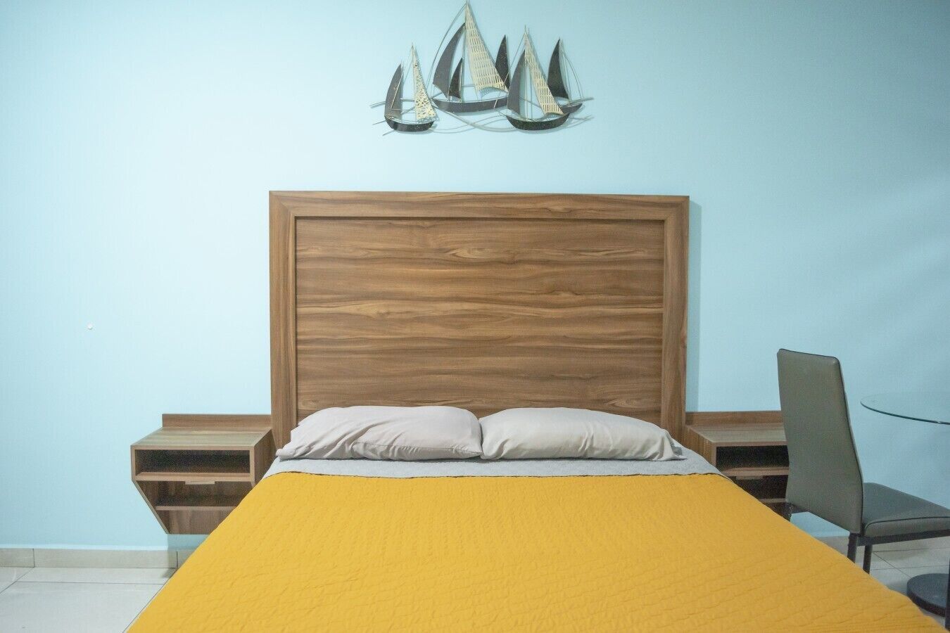 Ein Schlafzimmer mit einem hölzernen Kopfteil, einem Bett mit gelber Bettdecke, einem Nachttisch, einem Sessel und einem an der Wand befestigten dekorativen Segelboot.