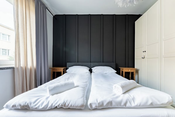 A double bed with white linens, a dark headboard, two bedside tables, and a window with curtains.