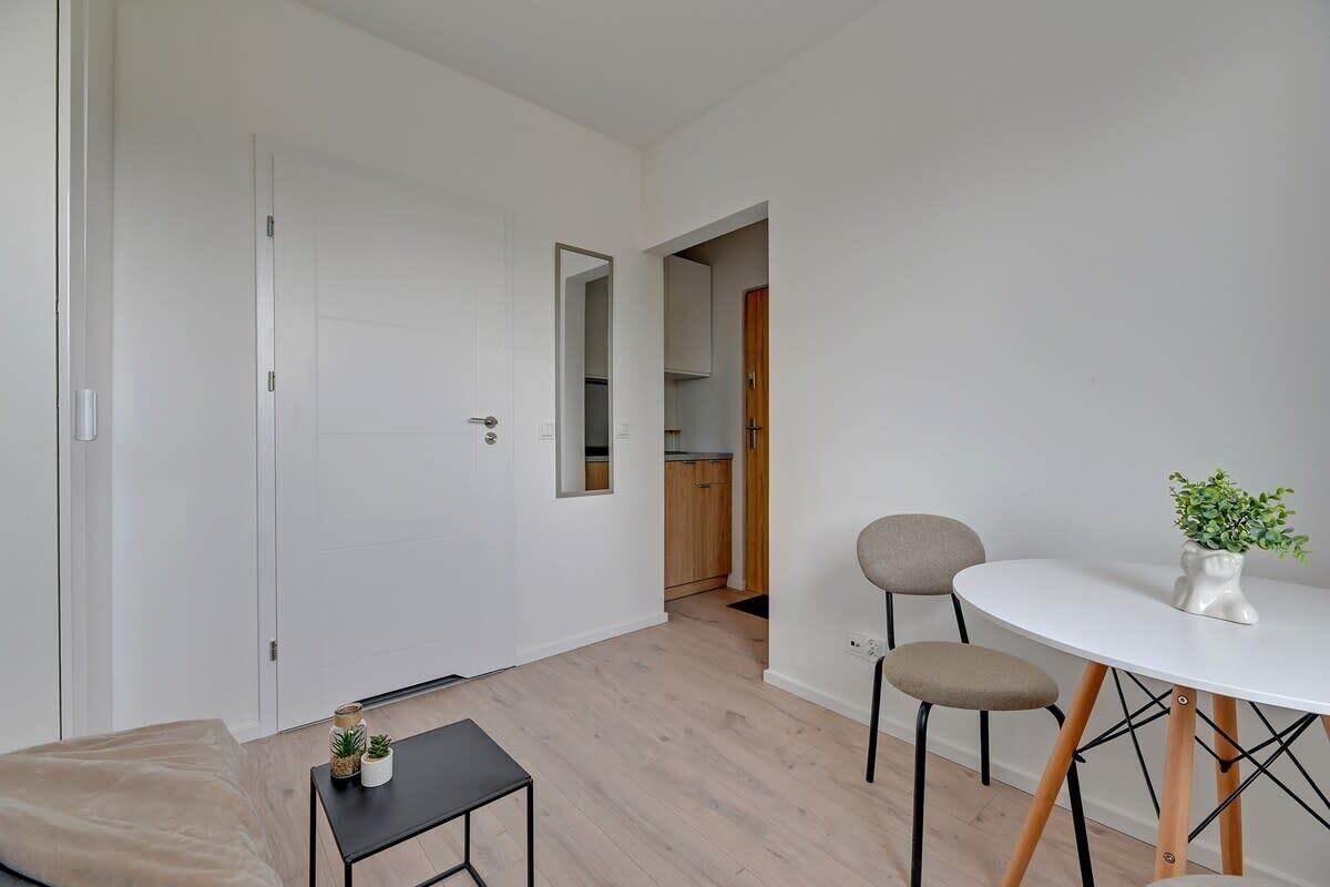Una habitación moderna con sofá, una mesita con una planta, una mesa redonda con sillas y una puerta que da a una cocina.