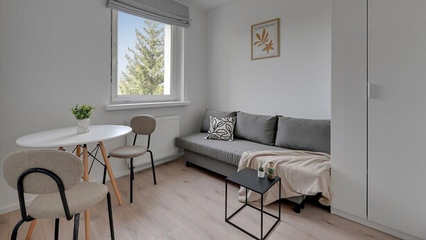 Een moderne woonkamer met een bank, een eettafel met stoelen, een bijzettafeltje en een raam met uitzicht op bomen.