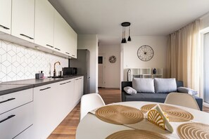 Una cocina moderna con armarios blancos, un comedor con mesa y sillas, una sala de estar con sofá y un reloj de pared grande.