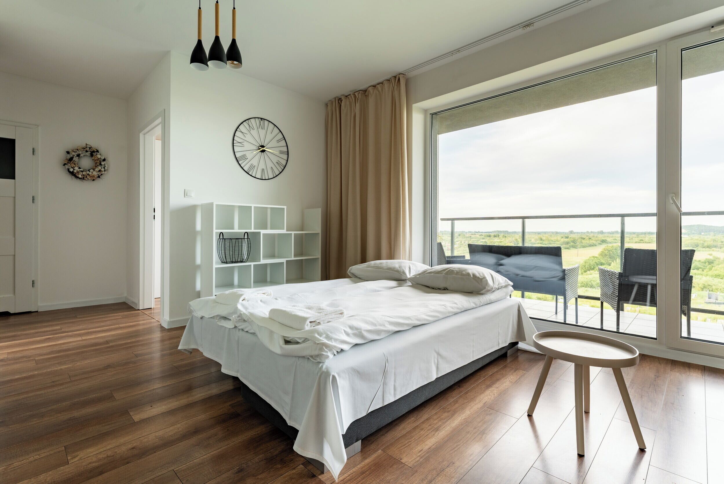 A bedroom with a bed, a round table, a large window with a view, a clock, and a shelf with white cabinets.