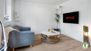 Moderni olohuone, jossa on harmaa sohva, pyöreä sohvapöytä, jossa on kultaisia yksityiskohtia, ruukkukasvi ja seinälle asennettu televisio, jonka näytöllä näkyy teksti ”NETFLIX”.
