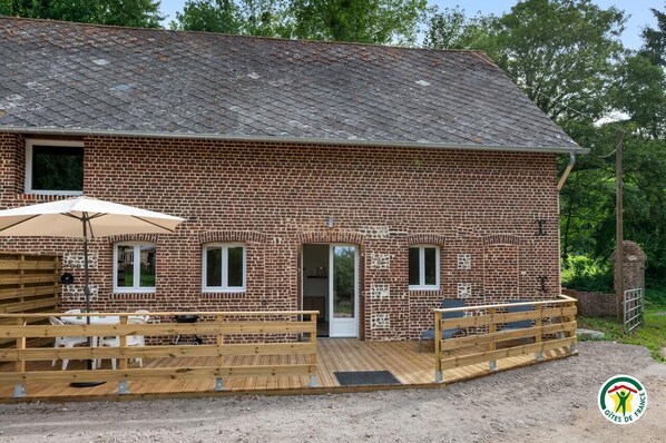 A brick building with a wooden deck, a door, and a window. - Gîtes de France® - L'Escapade (COTTEVRARD)