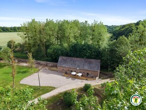 A single-story brick building with a flat roof, a paved driveway, and a small patio area with outdoor seating. - Gîtes de France® - L'Escapade (COTTEVRARD)