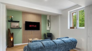 A modern living room with a flat-screen TV displaying 'NETFLIX,' a wooden accent wall, a sofa, a chair, and a window with a view.