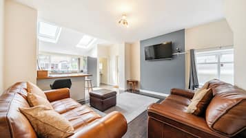 A modern living room with leather sofas, a flat-screen TV, and a kitchen in the background.