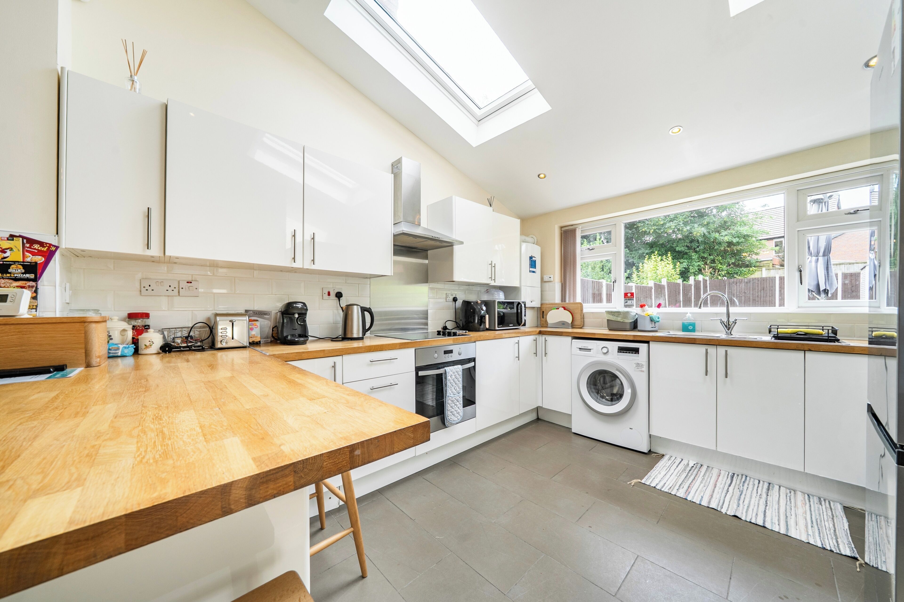A modern kitchen with white cabinets, a wooden countertop, a skylight, a washing machine, and a window with a view of the outside.