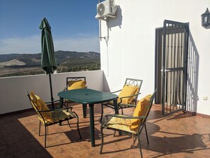 Eine Terrasse mit Tisch und Stühlen, einem grünen Sonnenschirm und Blick auf Hügel.