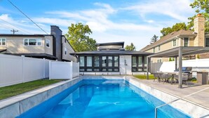 Una piscina rectangular con borde de azulejos, un patio cubierto con mobiliario exterior, y una valla blanca que rodea la propiedad.